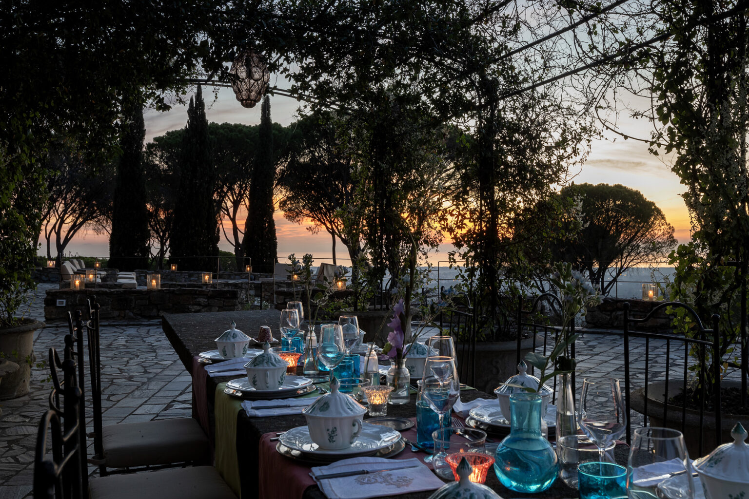 Elegant outdoor dining setup at sunset, featuring a beautifully arranged table with colorful glassware and fine dining essentials, surrounded by lush greenery and scenic views. | Only on SopranoVillas