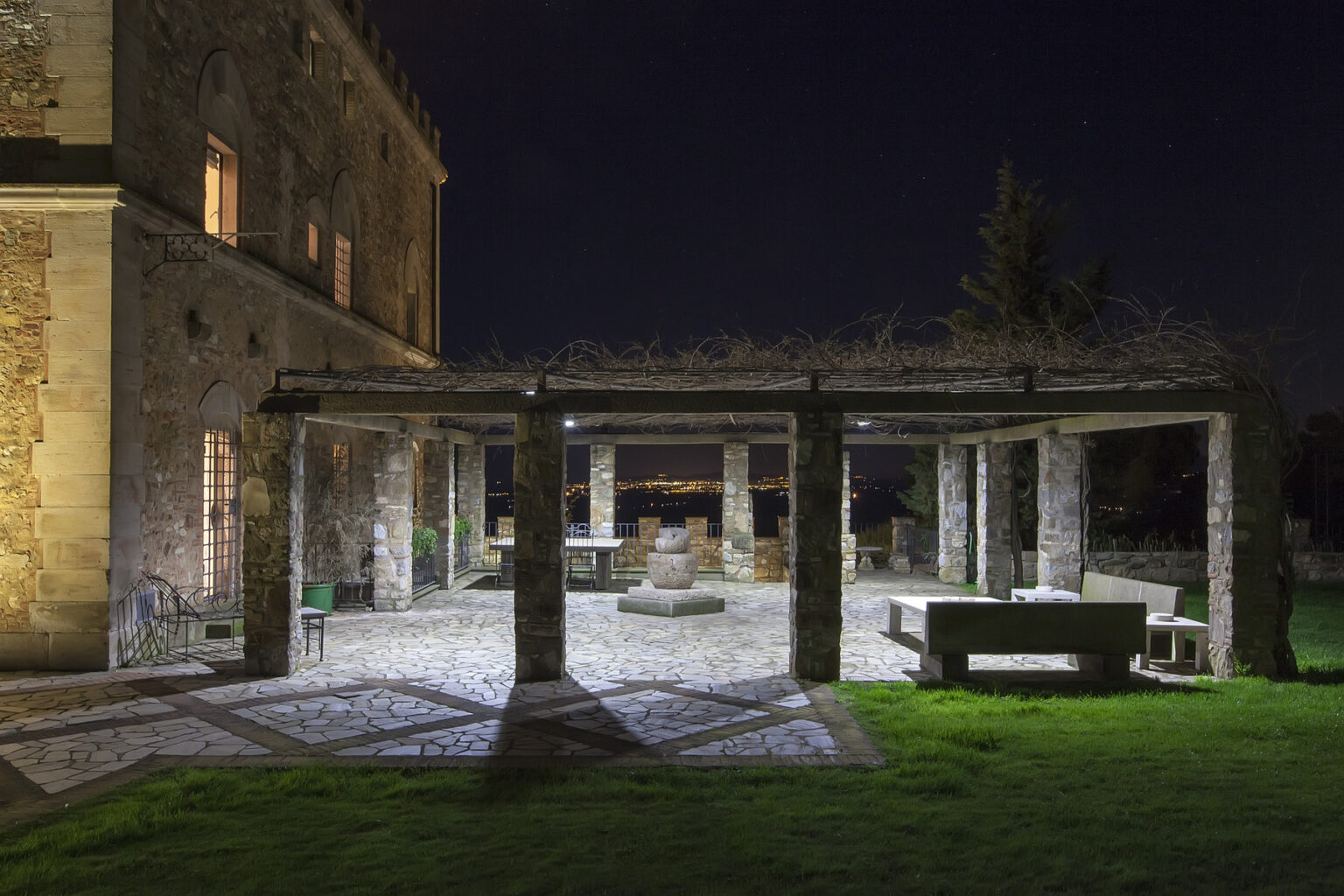 Nighttime view of a stone patio with a pergola, surrounded by greenery, featuring outdoor seating and a fire pit, near a historic building. | Only on SopranoVillas