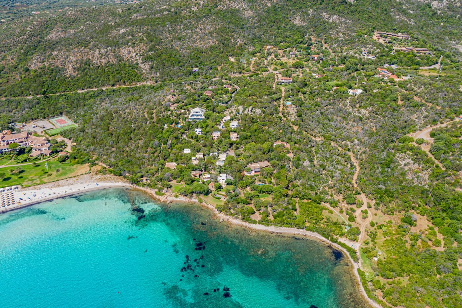 Aerial view of a coastal area featuring clear turquoise waters, sandy beach, and lush green hills with scattered residential buildings and pathways. | Only on SopranoVillas