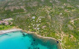 Aerial view of a coastal area featuring clear turquoise waters, sandy beach, and lush green hills with scattered residential buildings and pathways.