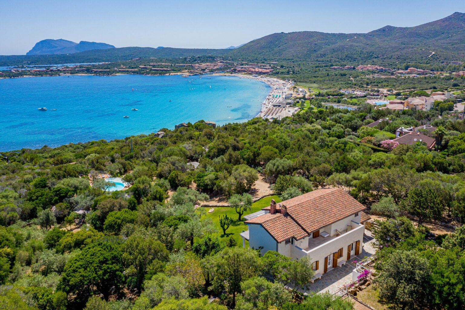 Aerial view of a picturesque coastal landscape featuring a white house surrounded by lush greenery, with a sandy beach and turquoise sea in the background, showcasing the natural beauty of the region. | Only on SopranoVillas