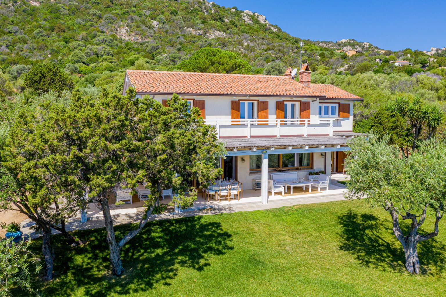 Aerial view of a charming two-story villa surrounded by lush greenery, featuring a spacious patio with outdoor seating and orange shutters, set against a backdrop of rolling hills. | Only on SopranoVillas