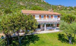 Aerial view of a charming two-story villa surrounded by lush greenery, featuring a spacious patio with outdoor seating and orange shutters, set against a backdrop of rolling hills.