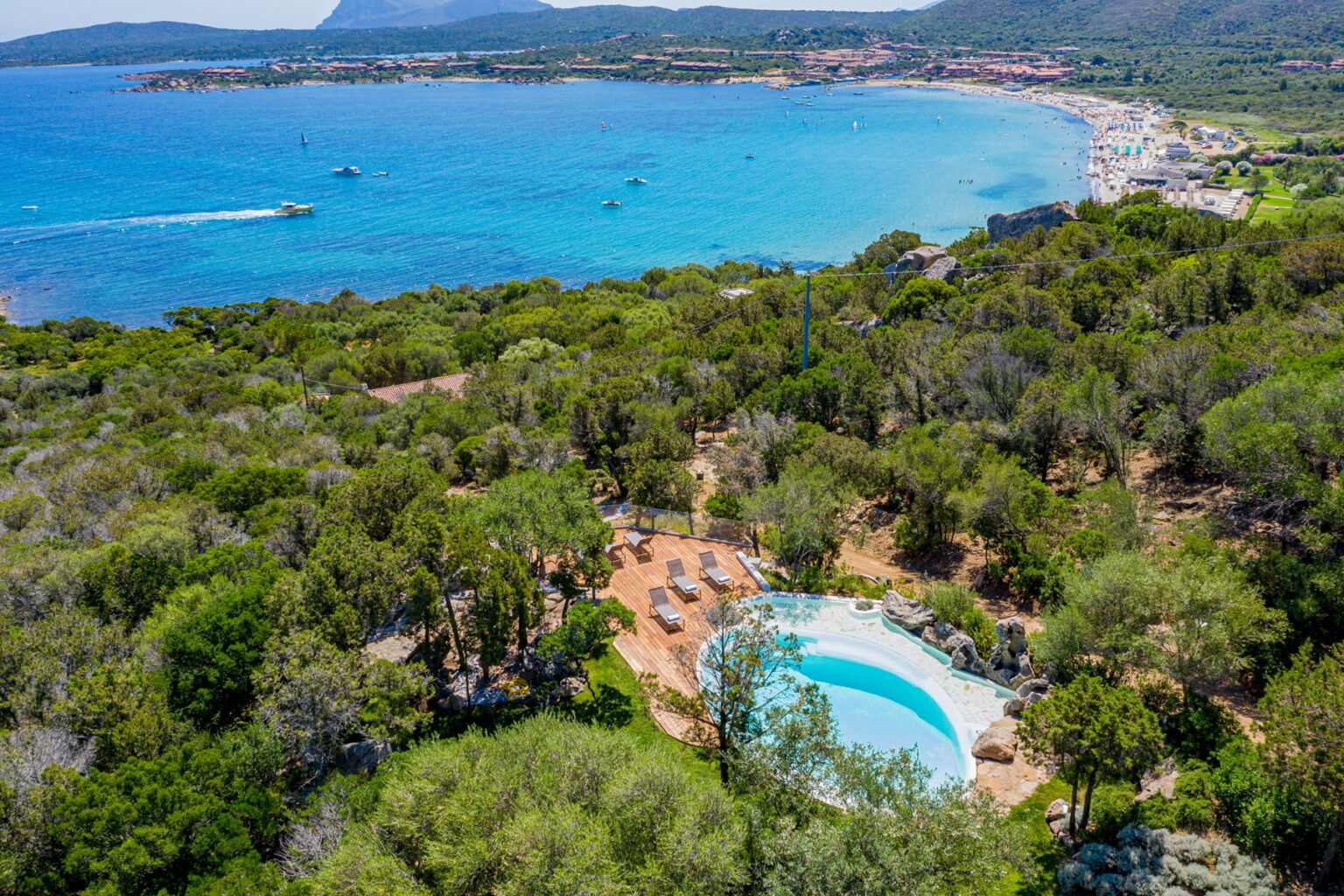 Aerial view of a serene coastal landscape featuring a swimming pool surrounded by lush greenery, with a clear blue sea and beach in the background, ideal for vacation and relaxation. | Only on SopranoVillas
