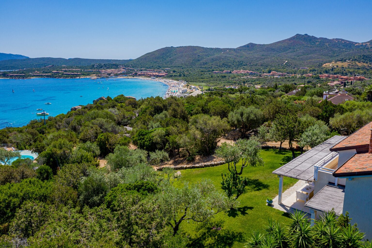Aerial view of a coastal landscape featuring lush greenery, a sandy beach, and distant mountains under a clear blue sky, showcasing a serene vacation destination. | Only on SopranoVillas