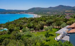 Aerial view of a coastal landscape featuring lush greenery, a sandy beach, and distant mountains under a clear blue sky, showcasing a serene vacation destination.