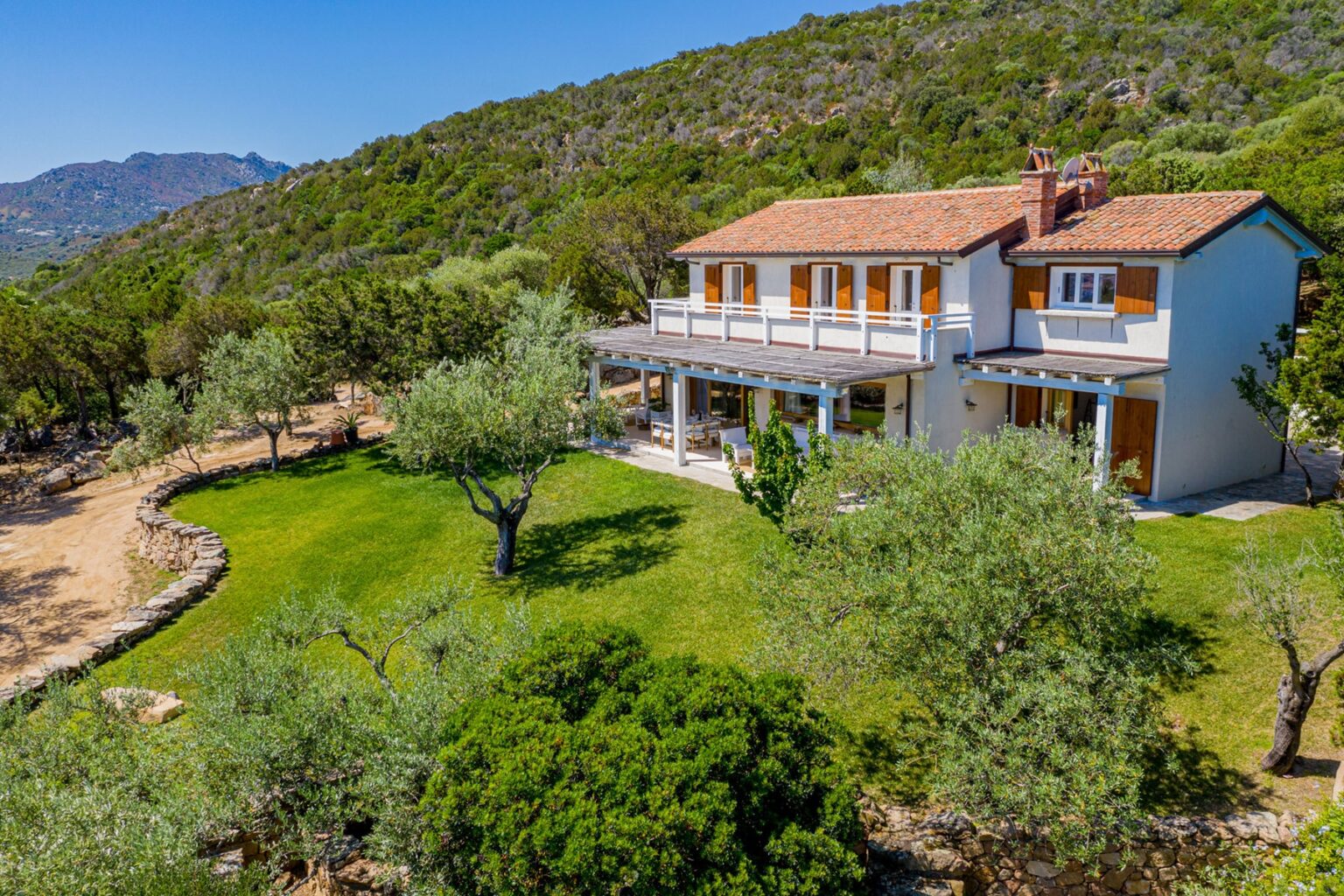 Aerial view of a charming two-story house surrounded by lush greenery and olive trees, with a spacious lawn and mountains in the background, showcasing a serene countryside setting. | Only on SopranoVillas