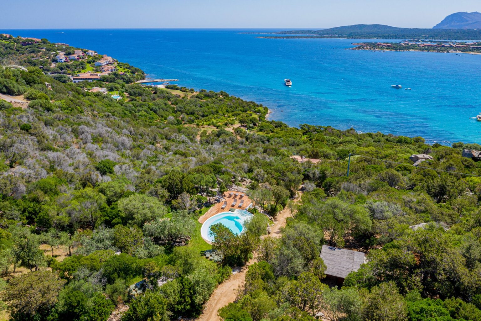 Aerial view of a lush green landscape featuring a swimming pool, surrounded by trees and a stunning blue ocean in the background, ideal for vacation rentals or coastal retreats. | Only on SopranoVillas