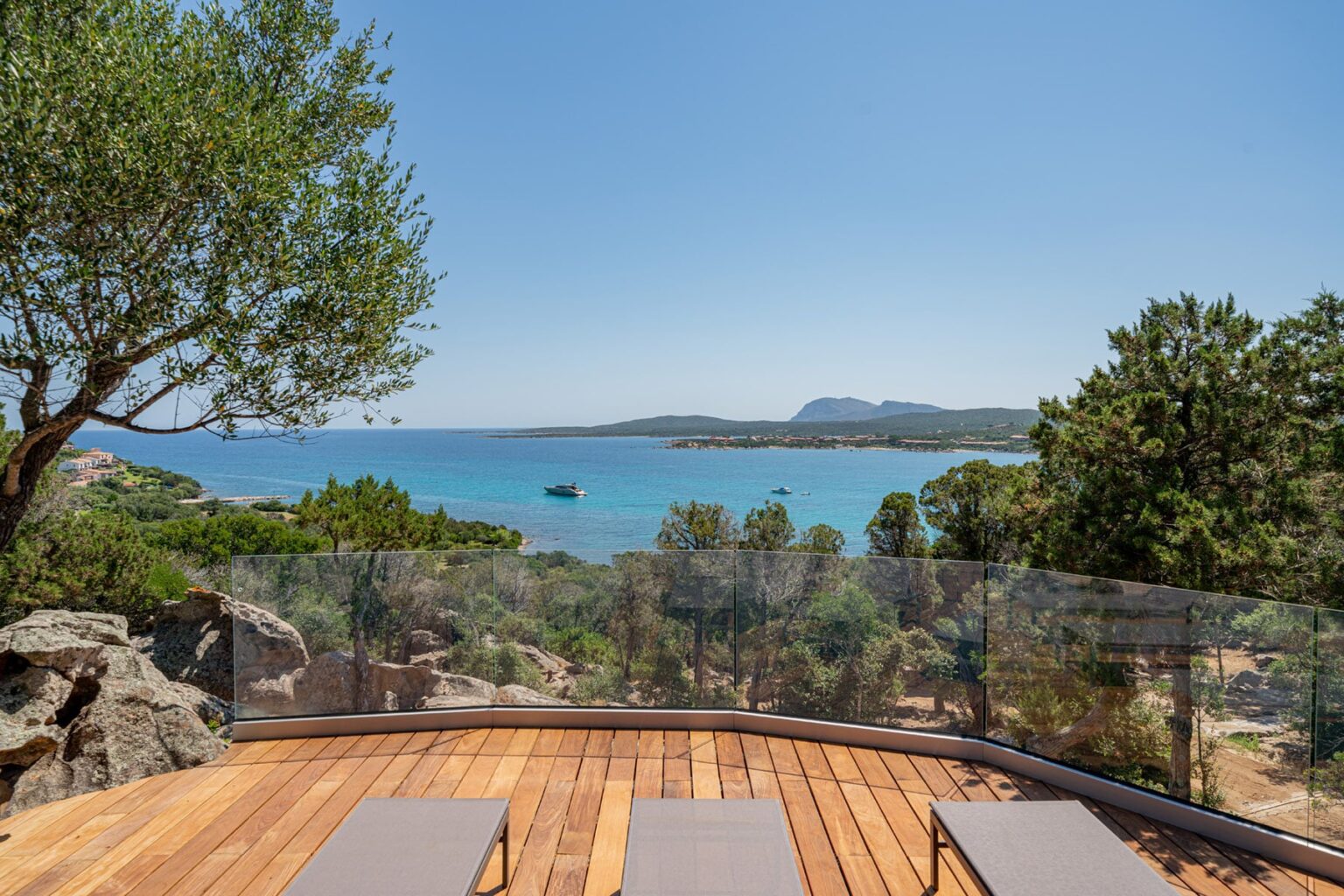 Scenic view of a tranquil beach from a wooden deck, featuring lounge chairs and lush greenery, with clear blue water and distant hills under a bright sky. Ideal for vacation and relaxation imagery. | Only on SopranoVillas