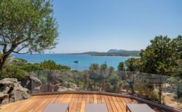 Scenic view of a tranquil beach from a wooden deck, featuring lounge chairs and lush greenery, with clear blue water and distant hills under a bright sky. Ideal for vacation and relaxation imagery.