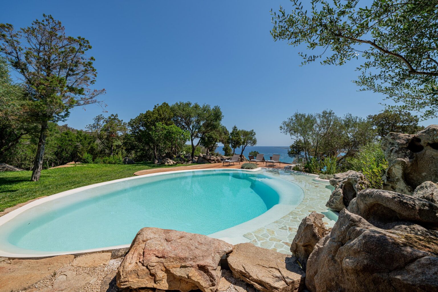 Scenic outdoor swimming pool surrounded by lush greenery and rocky landscape, with a clear blue sky and ocean view in the background. Ideal for relaxation and leisure activities. | Only on SopranoVillas