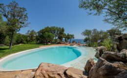 Scenic outdoor swimming pool surrounded by lush greenery and rocky landscape, with a clear blue sky and ocean view in the background. Ideal for relaxation and leisure activities.