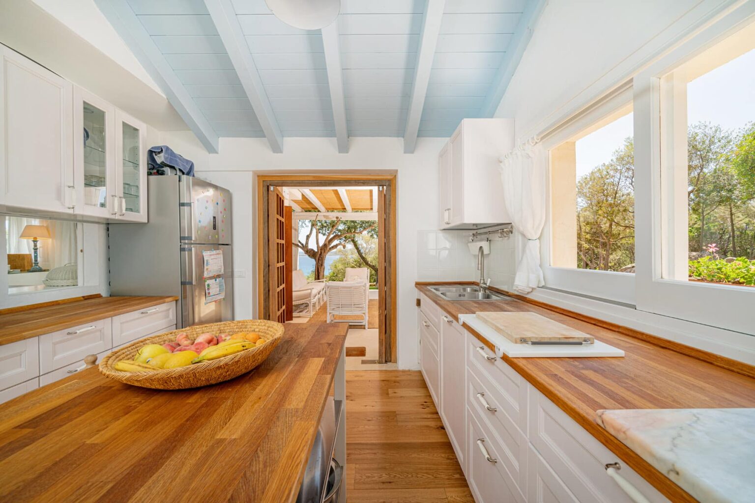 Bright and airy kitchen featuring wooden countertops, white cabinets, and large windows that open to a scenic outdoor view. A bowl of fresh fruits is displayed on the counter, enhancing the inviting atmosphere. | Only on SopranoVillas