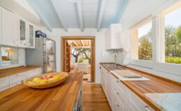 Bright and airy kitchen featuring wooden countertops, white cabinets, and large windows that open to a scenic outdoor view. A bowl of fresh fruits is displayed on the counter, enhancing the inviting atmosphere.