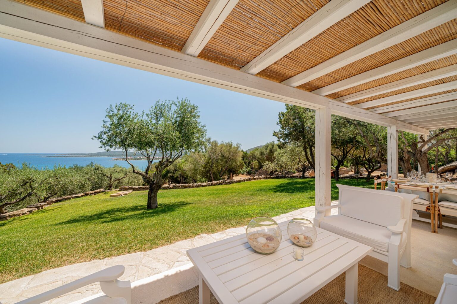 Scenic view from a terrace overlooking a lush green landscape with olive trees and the sea, featuring a white outdoor seating area and a sunny sky. | Only on SopranoVillas
