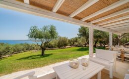 Scenic view from a terrace overlooking a lush green landscape with olive trees and the sea, featuring a white outdoor seating area and a sunny sky.