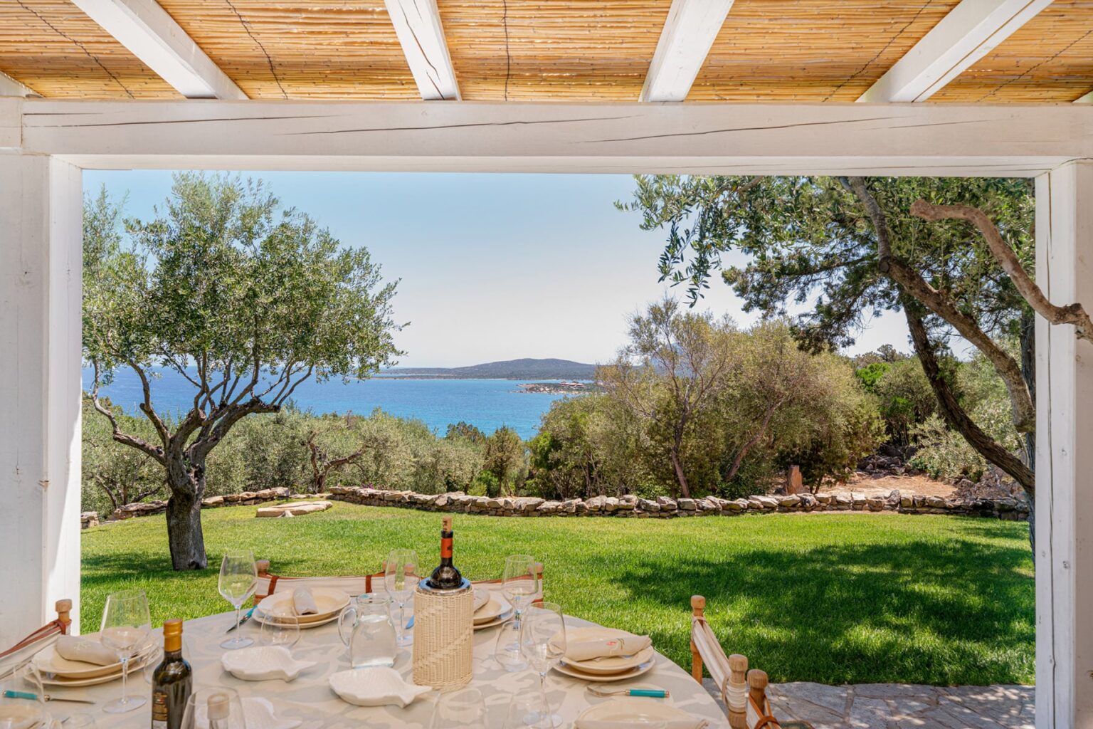 Scenic view from a dining area overlooking a tranquil sea, surrounded by lush greenery and olive trees, perfect for outdoor gatherings. | Only on SopranoVillas