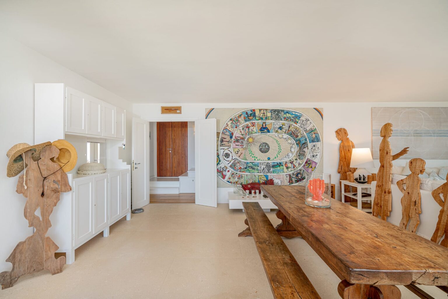 Modern interior design featuring a spacious dining area with a large wooden table, unique wooden sculptures, and a colorful wall mural. Bright, airy space with white cabinetry and natural light. Ideal for showcasing contemporary art and rustic decor elements. | Only on SopranoVillas