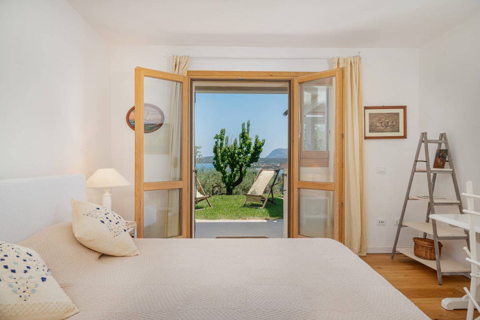 Bright and airy bedroom with a large open window showcasing a scenic view of a garden and mountains, featuring a comfortable bed with decorative pillows, a cozy lamp, and a stylish ladder shelf. | Only on SopranoVillas