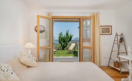 Bright and airy bedroom with a large open window showcasing a scenic view of a garden and mountains, featuring a comfortable bed with decorative pillows, a cozy lamp, and a stylish ladder shelf.