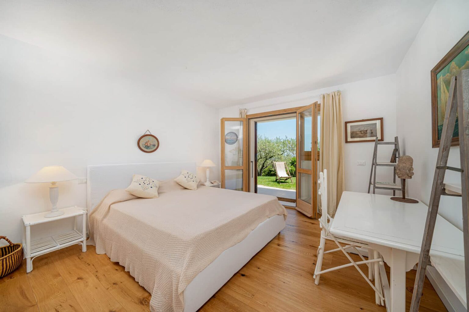 Bright and airy bedroom featuring a queen-sized bed with neutral bedding, white nightstands, and a desk. Large doors open to a scenic outdoor view, complemented by natural light and wooden flooring, creating a serene atmosphere. | Only on SopranoVillas