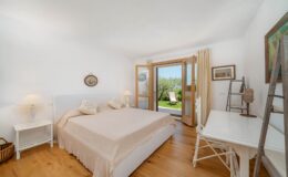 Bright and airy bedroom featuring a queen-sized bed with neutral bedding, white nightstands, and a desk. Large doors open to a scenic outdoor view, complemented by natural light and wooden flooring, creating a serene atmosphere.