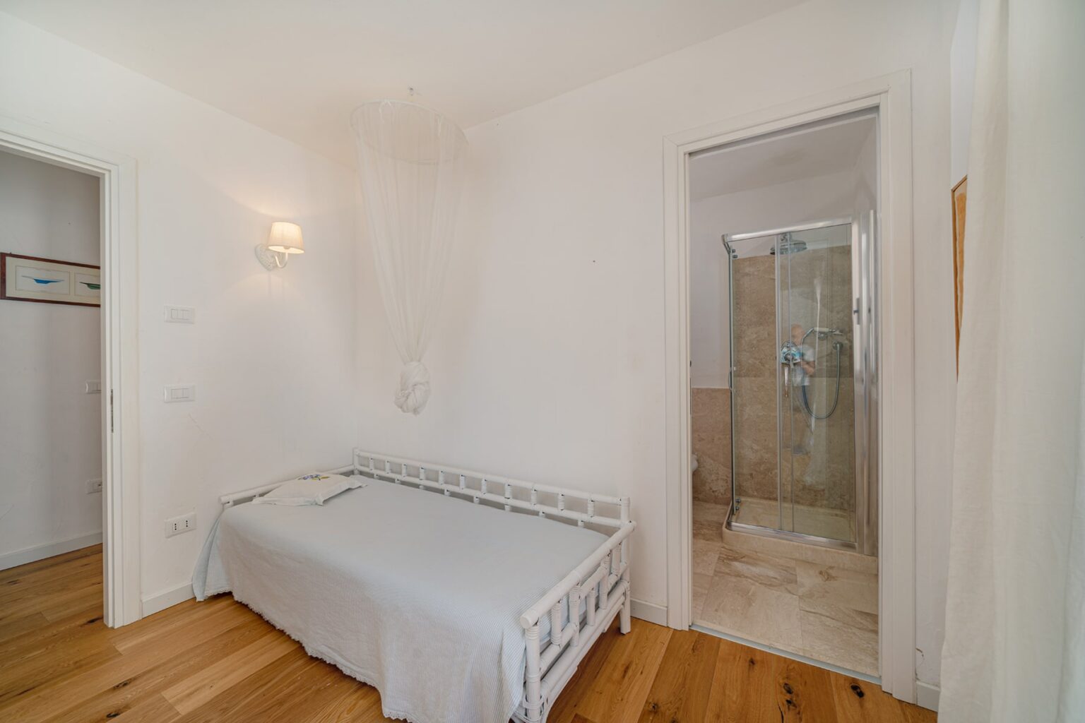 Bright and modern bedroom featuring a white bed, wooden flooring, and an en-suite shower room with glass doors. The minimalist decor includes soft lighting and a serene atmosphere, ideal for relaxation. | Only on SopranoVillas