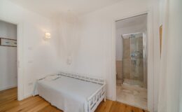 Bright and modern bedroom featuring a white bed, wooden flooring, and an en-suite shower room with glass doors. The minimalist decor includes soft lighting and a serene atmosphere, ideal for relaxation.