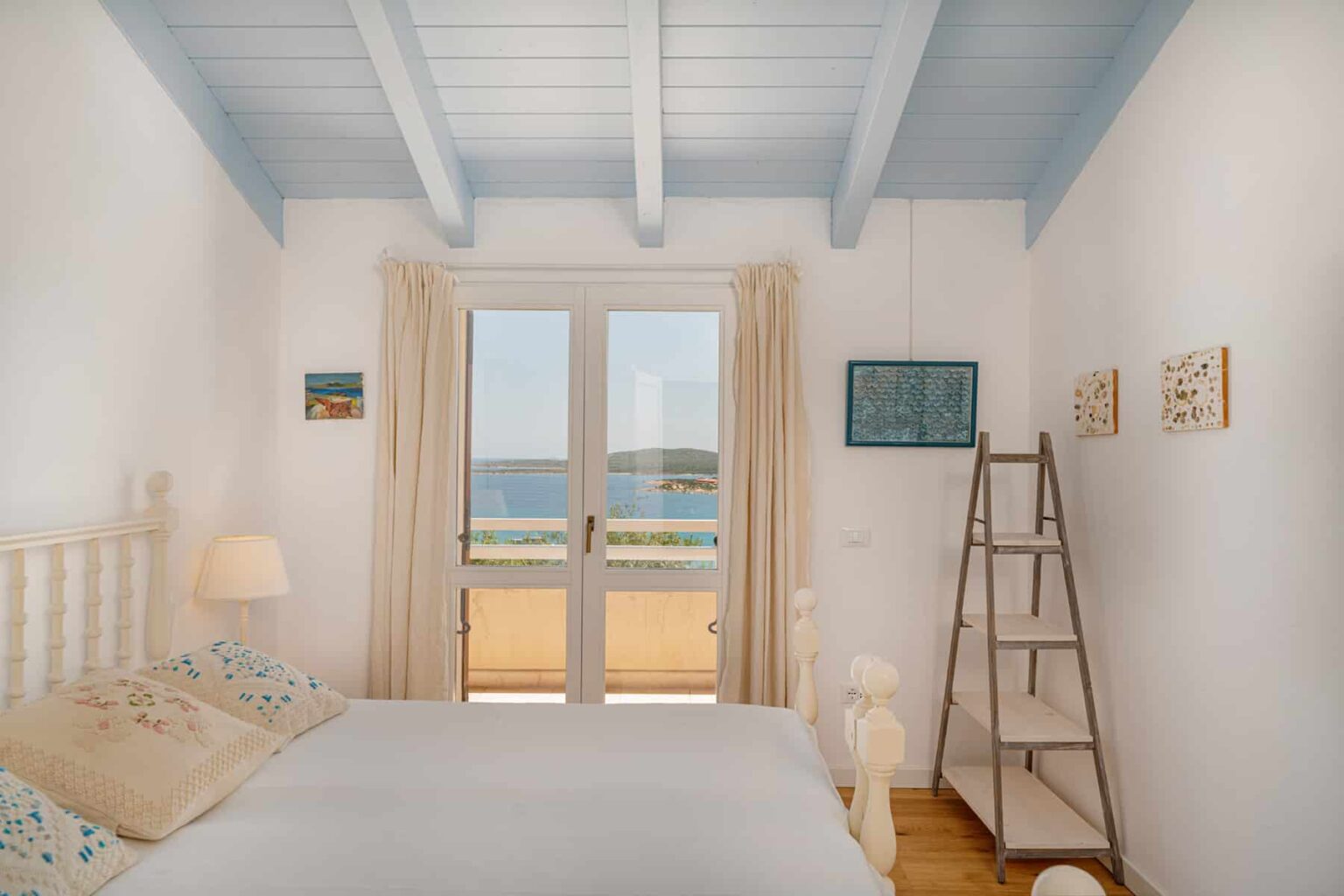 Cozy bedroom with white wooden furniture, blue ceiling beams, and large windows offering a scenic view of the sea. The room features soft lighting, decorative wall art, and a wooden ladder, creating a serene and inviting atmosphere. | Only on SopranoVillas