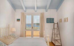 Cozy bedroom with white wooden furniture, blue ceiling beams, and large windows offering a scenic view of the sea. The room features soft lighting, decorative wall art, and a wooden ladder, creating a serene and inviting atmosphere.