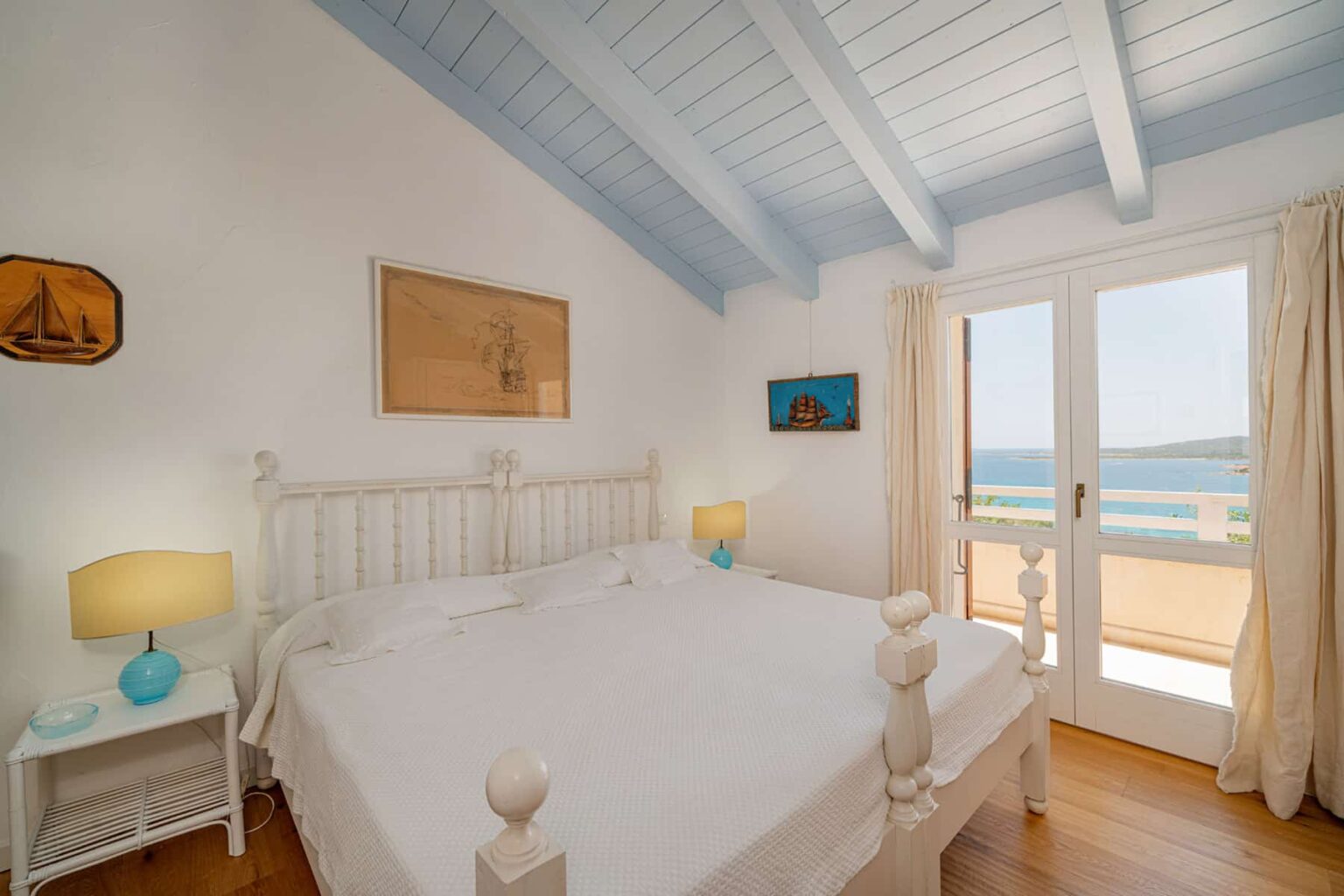 Bright and airy bedroom with a white wooden bed, blue ceiling beams, and natural light streaming through large glass doors leading to a scenic view. The room features light-colored walls, a nightstand with a turquoise lamp, and coastal artwork, creating a serene atmosphere. | Only on SopranoVillas