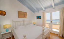 Bright and airy bedroom with a white wooden bed, blue ceiling beams, and natural light streaming through large glass doors leading to a scenic view. The room features light-colored walls, a nightstand with a turquoise lamp, and coastal artwork, creating a serene atmosphere.