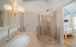 Modern bathroom interior featuring a glass shower enclosure, marble countertop, and elegant lighting fixtures. The design showcases a clean, spacious layout with natural light streaming in from an adjacent room.