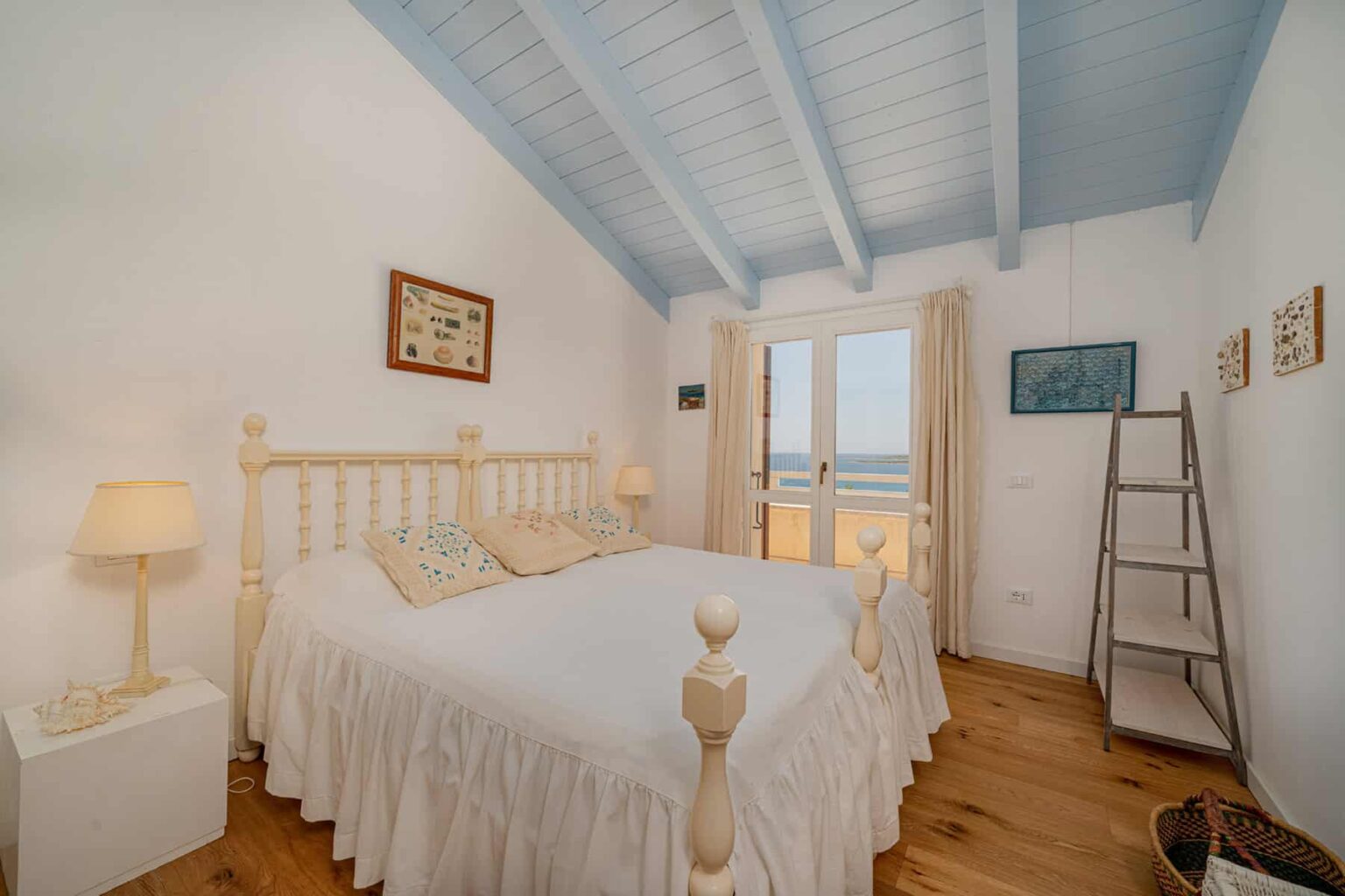 Cozy bedroom with a white wooden bed, featuring soft bedding and decorative pillows, complemented by natural light from large windows. The room has a light blue ceiling, wooden flooring, and minimalist decor, creating a serene and inviting atmosphere. | Only on SopranoVillas