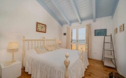 Cozy bedroom with a white wooden bed, featuring soft bedding and decorative pillows, complemented by natural light from large windows. The room has a light blue ceiling, wooden flooring, and minimalist decor, creating a serene and inviting atmosphere.