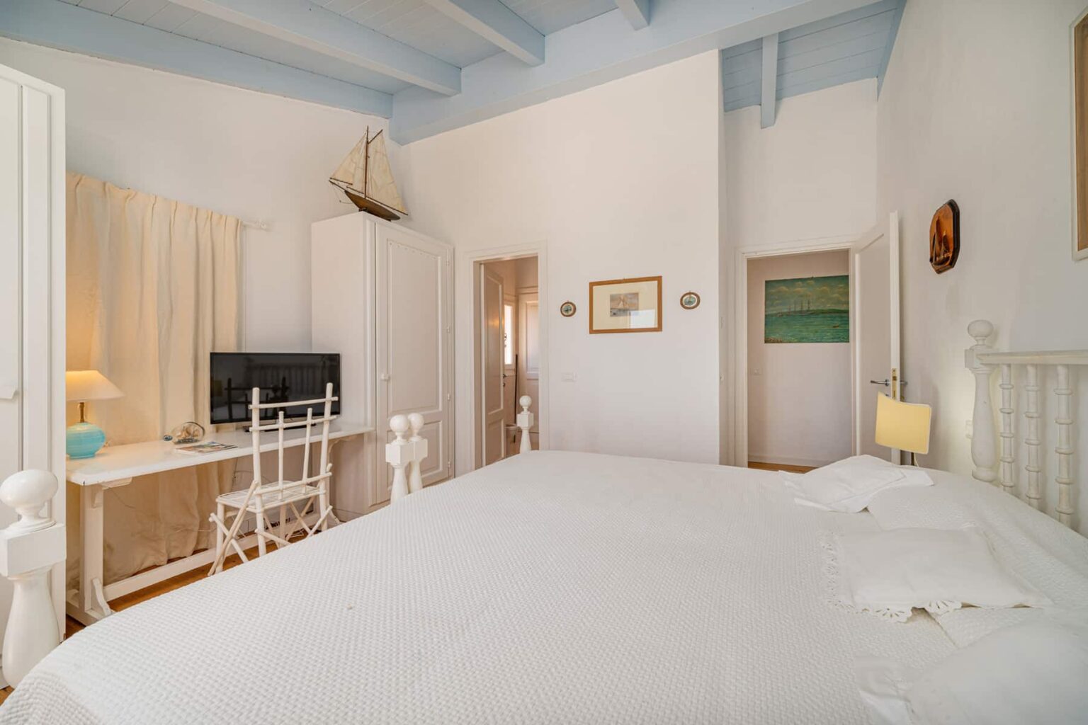 Bright and airy bedroom featuring a large bed, a desk with a chair, and a television. The room has white walls, blue ceiling beams, and minimalistic decor, creating a serene atmosphere. | Only on SopranoVillas