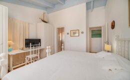 Bright and airy bedroom featuring a large bed, a desk with a chair, and a television. The room has white walls, blue ceiling beams, and minimalistic decor, creating a serene atmosphere.
