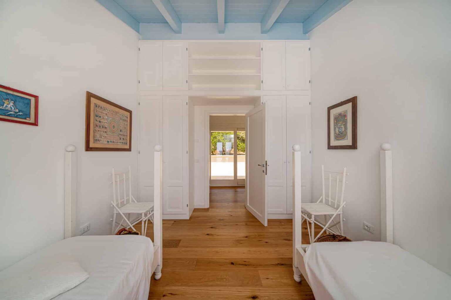 Bright and airy bedroom featuring two white twin beds, wooden flooring, and blue ceiling beams. The room includes framed artwork on the walls and a doorway leading to a sunlit area. Ideal for a cozy and relaxing stay. | Only on SopranoVillas