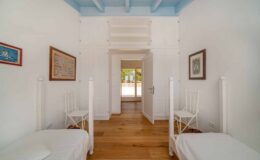 Bright and airy bedroom featuring two white twin beds, wooden flooring, and blue ceiling beams. The room includes framed artwork on the walls and a doorway leading to a sunlit area. Ideal for a cozy and relaxing stay.