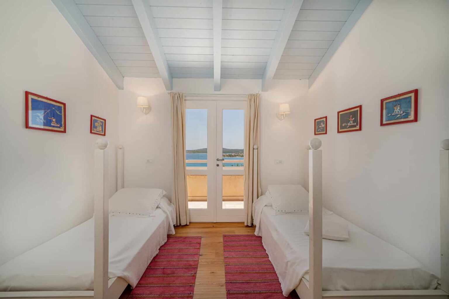 Bright and airy bedroom featuring two twin beds, white walls, and wooden beams, with large windows offering a view of the sea. Decorated with colorful artwork and a cozy rug, this space is perfect for relaxation. | Only on SopranoVillas