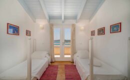 Bright and airy bedroom featuring two twin beds, white walls, and wooden beams, with large windows offering a view of the sea. Decorated with colorful artwork and a cozy rug, this space is perfect for relaxation.