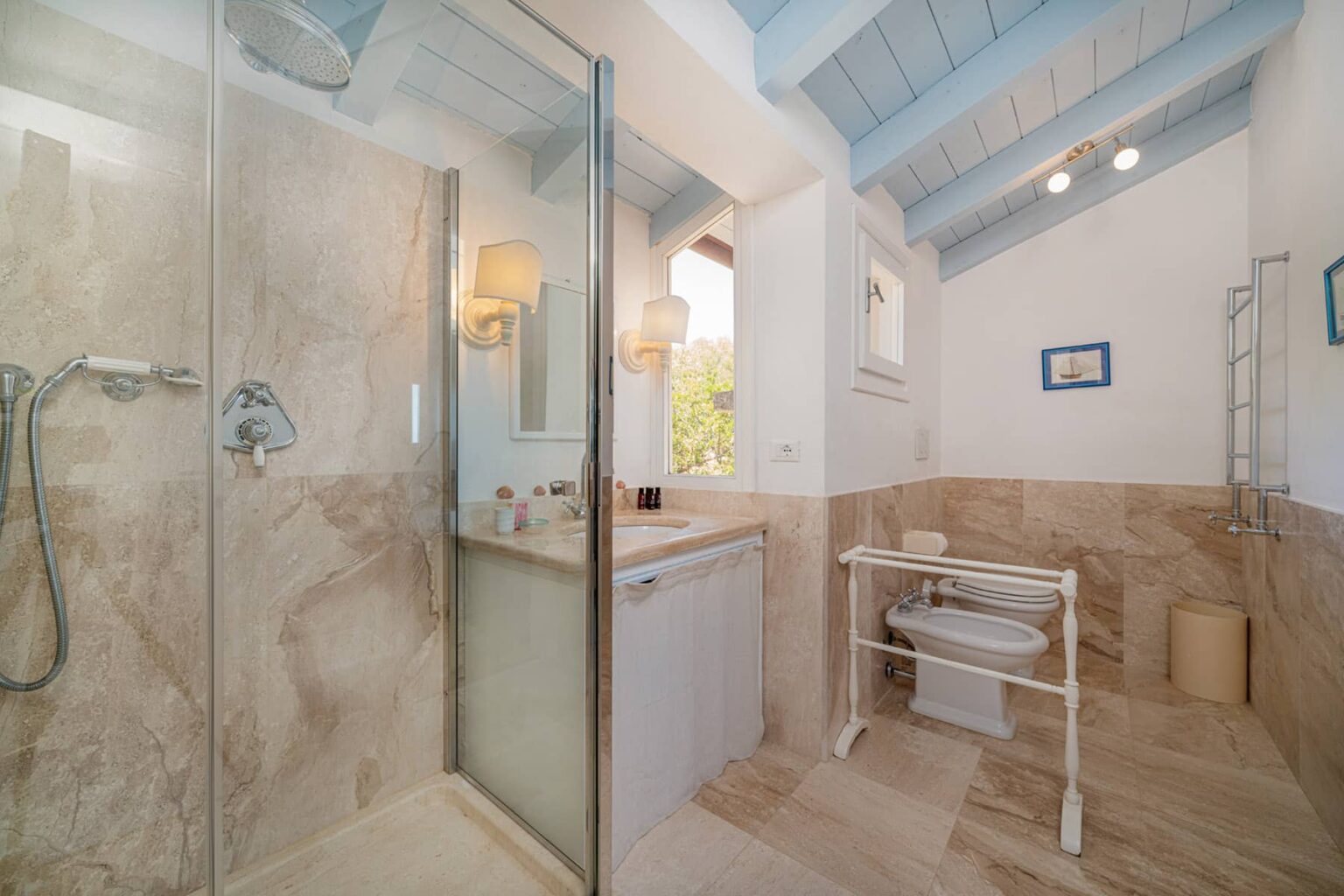 Modern bathroom interior featuring a glass shower enclosure, marble tiles, and a stylish sink area with natural lighting. The ceiling has a light blue finish, adding a touch of elegance. | Only on SopranoVillas