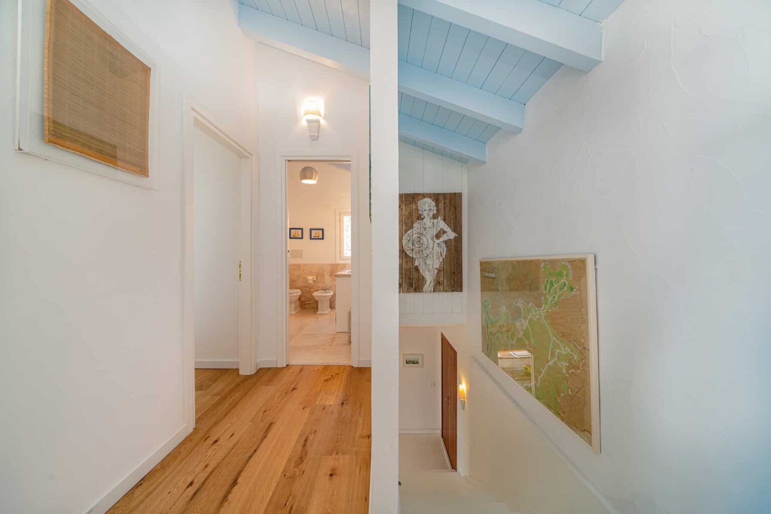 Bright and airy hallway featuring light wood flooring, white walls, and blue ceiling beams. Artwork adorns the walls, enhancing the modern aesthetic. The open layout leads to a cozy living space, showcasing a blend of style and comfort. | Only on SopranoVillas