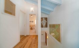 Bright and airy hallway featuring light wood flooring, white walls, and blue ceiling beams. Artwork adorns the walls, enhancing the modern aesthetic. The open layout leads to a cozy living space, showcasing a blend of style and comfort.