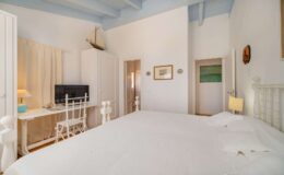 Bright and airy bedroom featuring a large white bed, a desk with a chair, and a television. The room has light blue beams and soft, neutral decor, creating a serene atmosphere. Ideal for relaxation and comfort.