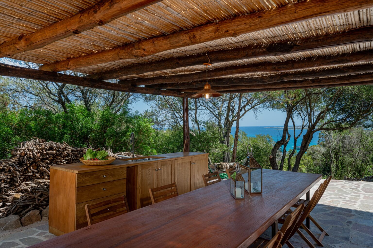 Outdoor dining area with a wooden table and chairs under a thatched roof, surrounded by trees and a view of the ocean in the background. Ideal for relaxing meals in a serene setting. | Only on SopranoVillas