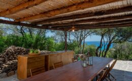 Outdoor dining area with a wooden table and chairs under a thatched roof, surrounded by trees and a view of the ocean in the background. Ideal for relaxing meals in a serene setting.