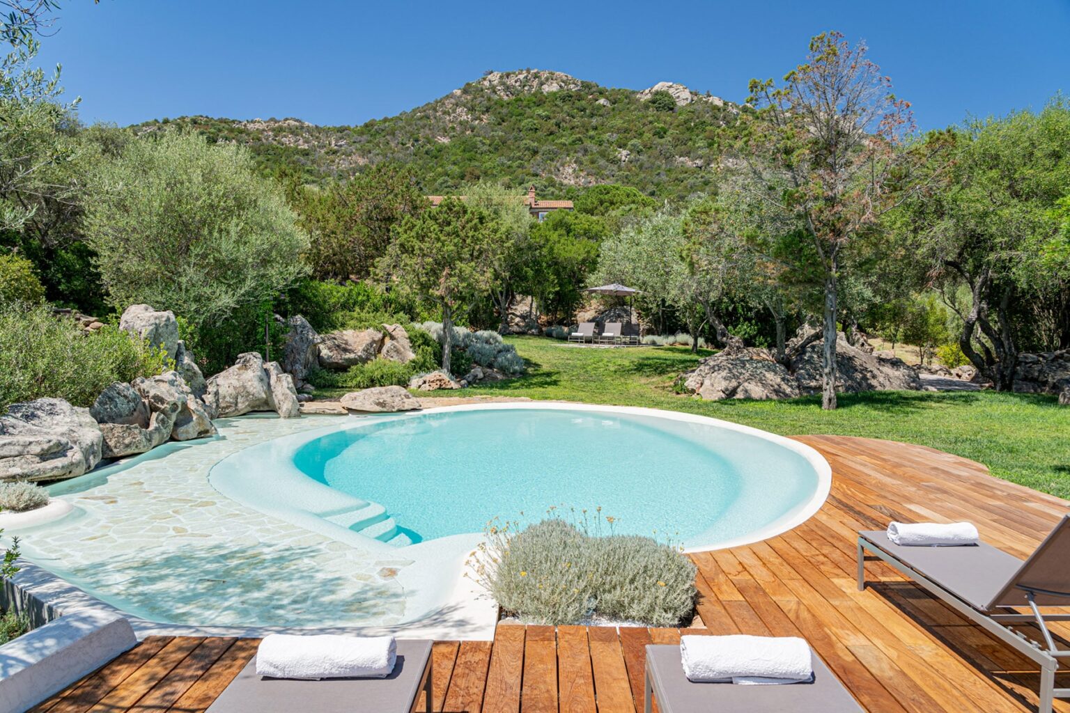 Serene outdoor swimming pool surrounded by lush greenery and rocky landscape, featuring wooden decking and lounge chairs, ideal for relaxation in a tranquil setting. | Only on SopranoVillas