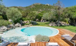Serene outdoor swimming pool surrounded by lush greenery and rocky landscape, featuring wooden decking and lounge chairs, ideal for relaxation in a tranquil setting.