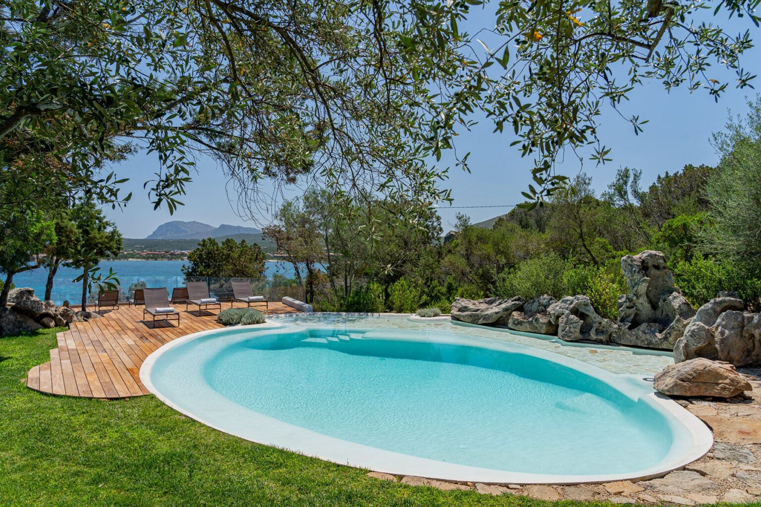 Serene outdoor swimming pool surrounded by lush greenery and natural rock formations, with a picturesque view of the water and mountains in the background. Ideal for relaxation and leisure. | Only on SopranoVillas